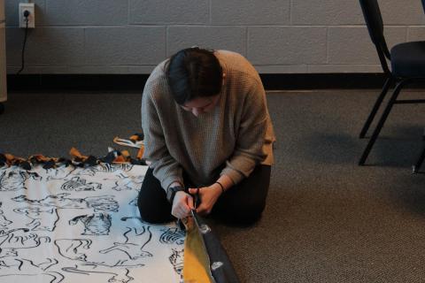 Student kneeling on floor cutting fleece fabric piece.